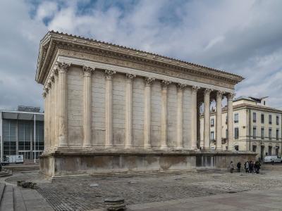 A low-angle photograph of the Maison Carrée, a rectangular Roman temple with Corinthian columns and a high podium, situated in a paved public square with modern buildings and a cloudy sky in the background.