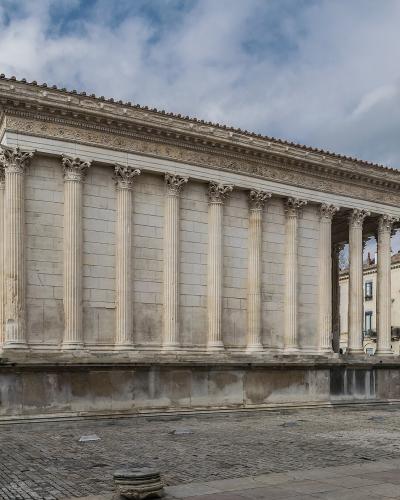 A low-angle photograph of the Maison Carrée, a rectangular Roman temple with Corinthian columns and a high podium, situated in a paved public square with modern buildings and a cloudy sky in the background.