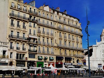 A street-level view of a curved, multi-story beige building with wrought-iron balconies and ground-floor shops including "DESSANGE" and "RESTAURANT," adjacent to a white church facade under a clear blue sky.