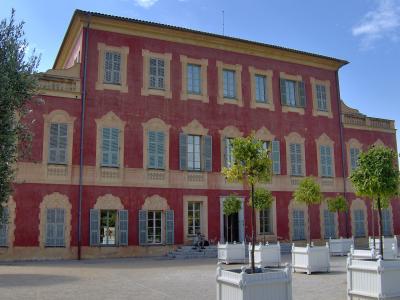 A large, three-story pink villa with yellow stone trim and blue shutters, featuring a courtyard with white planters and young trees in the foreground.