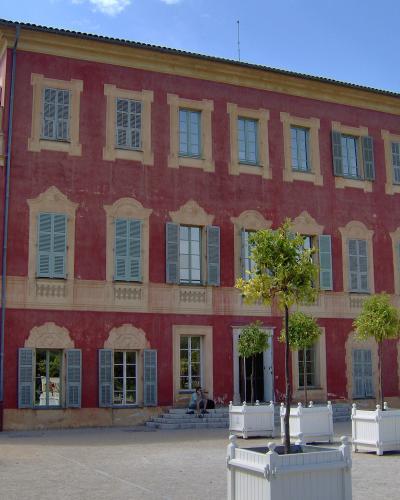 A large, three-story pink villa with yellow stone trim and blue shutters, featuring a courtyard with white planters and young trees in the foreground.