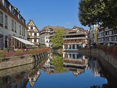 A scenic view of the Lauch River in the Petite France district of Strasbourg, France, featuring traditional half-timbered houses including the prominent Maison des Tanneurs on the right bank.