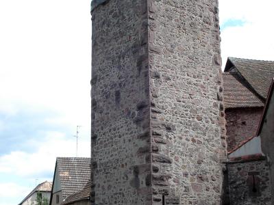 A tall, rectangular medieval stone tower with a pyramidal tiled roof, situated in a village setting with adjacent buildings and parked cars.