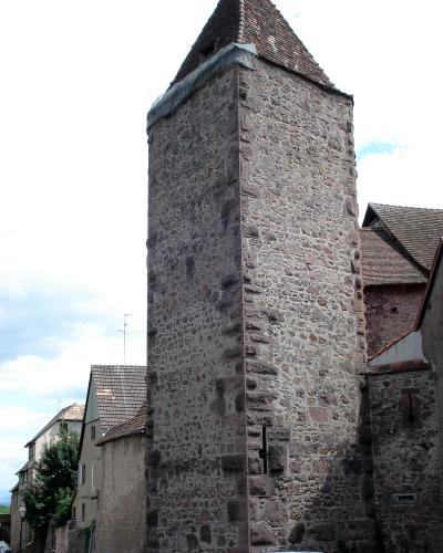 A tall, rectangular medieval stone tower with a pyramidal tiled roof, situated in a village setting with adjacent buildings and parked cars.