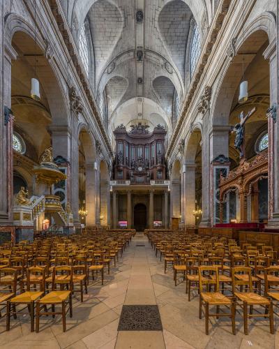 The image shows the interior nave of a large, historic church with tall fluted columns, arched ceilings, rows of wooden chairs, and stained-glass windows; no visible signage or text is present.