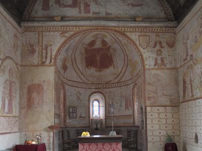 The image shows the interior of a historic church with extensive medieval frescoes on the walls and apse, a wooden altar covered with a red patterned cloth, a small stained-glass window above the altar, and a vaulted ceiling; no visible signage or