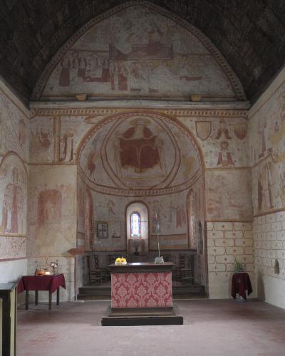 The image shows the interior of a historic church with extensive medieval frescoes on the walls and apse, a wooden altar covered with a red patterned cloth, a small stained-glass window above the altar, and a vaulted ceiling; no visible signage or