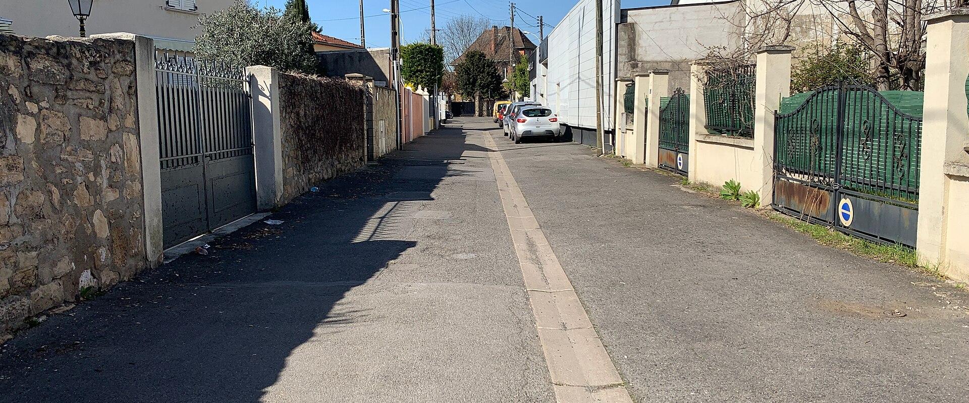 A narrow paved residential street in a French town, flanked by stone walls, houses with shutters, and green metal gates, under a clear blue sky; a white car is parked on the right side, and utility poles line the road.
