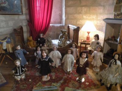 A display of vintage dolls and teddy bears arranged in a bedroom diorama within a stone-walled room, featuring period furniture, a red curtain, and a lamp.