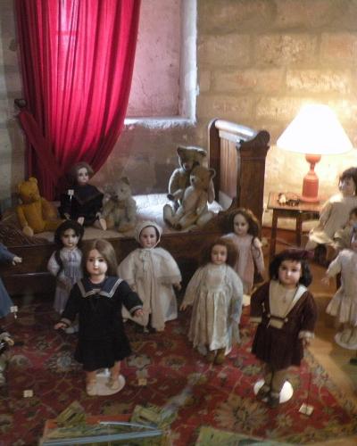 A display of vintage dolls and teddy bears arranged in a bedroom diorama within a stone-walled room, featuring period furniture, a red curtain, and a lamp.
