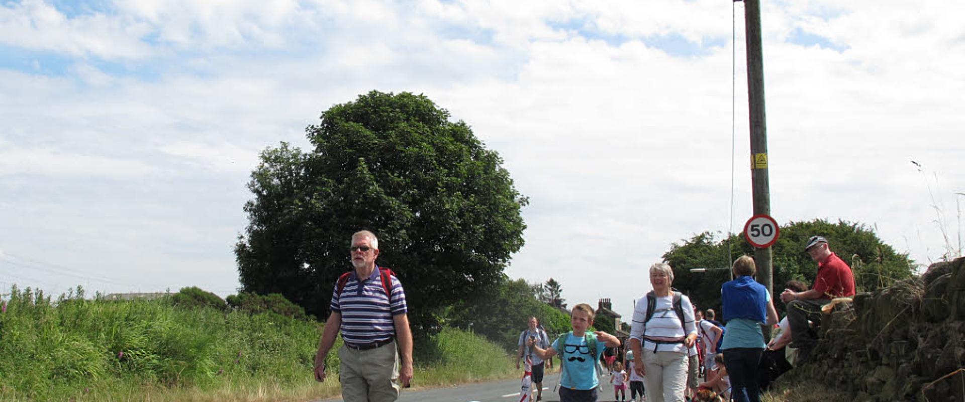A group of people walking along a paved road in a rural setting, with a man in a striped shirt and shorts in the foreground, a child holding an umbrella, and a stone wall on the right; a circular speed limit sign showing "50" is visible on a lampp