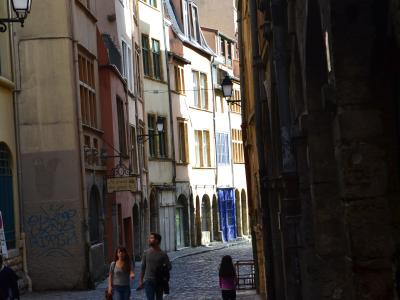 A narrow, cobblestone street in a historic European city, flanked by tall, multi-story buildings with Renaissance-style architecture, including arched doorways and shuttered windows; pedestrians walk along the street, and a sign reading "Au pavill