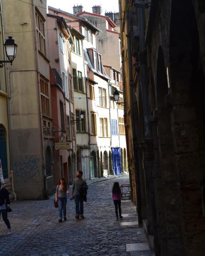 A narrow, cobblestone street in a historic European city, flanked by tall, multi-story buildings with Renaissance-style architecture, including arched doorways and shuttered windows; pedestrians walk along the street, and a sign reading "Au pavill
