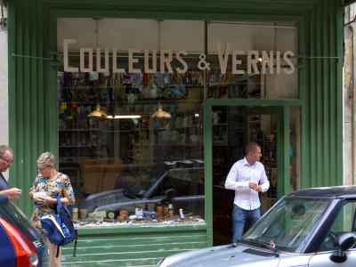 A street-level view of a green-painted storefront with a sign reading "Droguerie des - Halles" and window text "COULEURS & VERNIS", featuring pedestrians and parked cars on a paved sidewalk.