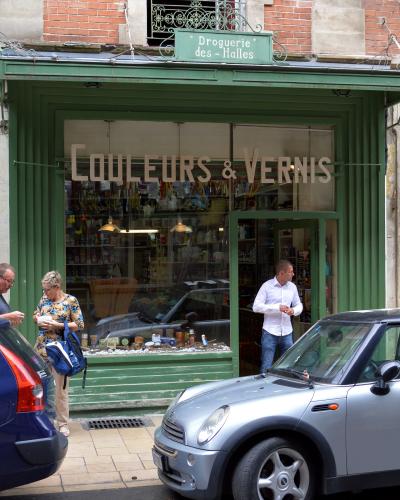 A street-level view of a green-painted storefront with a sign reading "Droguerie des - Halles" and window text "COULEURS & VERNIS", featuring pedestrians and parked cars on a paved sidewalk.