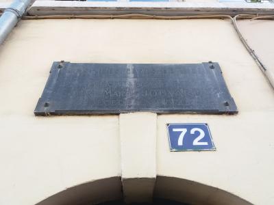 A close-up of a dark metal plaque mounted on a cream-colored wall, inscribed with "HOSPICES CIVILS DE LYON" and other text, positioned above a blue house number plate reading "72".