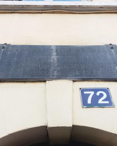 A close-up of a dark metal plaque mounted on a cream-colored wall, inscribed with "HOSPICES CIVILS DE LYON" and other text, positioned above a blue house number plate reading "72".