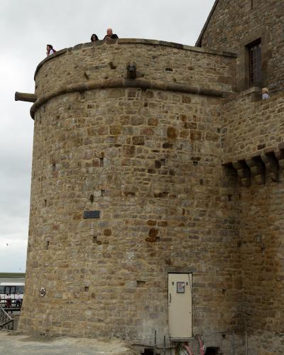 A stone tower with people on top, part of a larger stone building complex, under an overcast sky, with a bus visible in the lower left background.