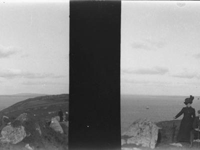 A black-and-white stereoscopic photograph showing three people (a woman standing and two men seated) on a grassy, rocky cliff edge overlooking the sea, with a distant coastline visible across the water.