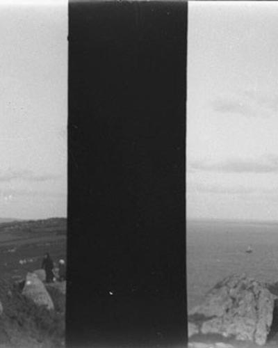A black-and-white stereoscopic photograph showing three people (a woman standing and two men seated) on a grassy, rocky cliff edge overlooking the sea, with a distant coastline visible across the water.