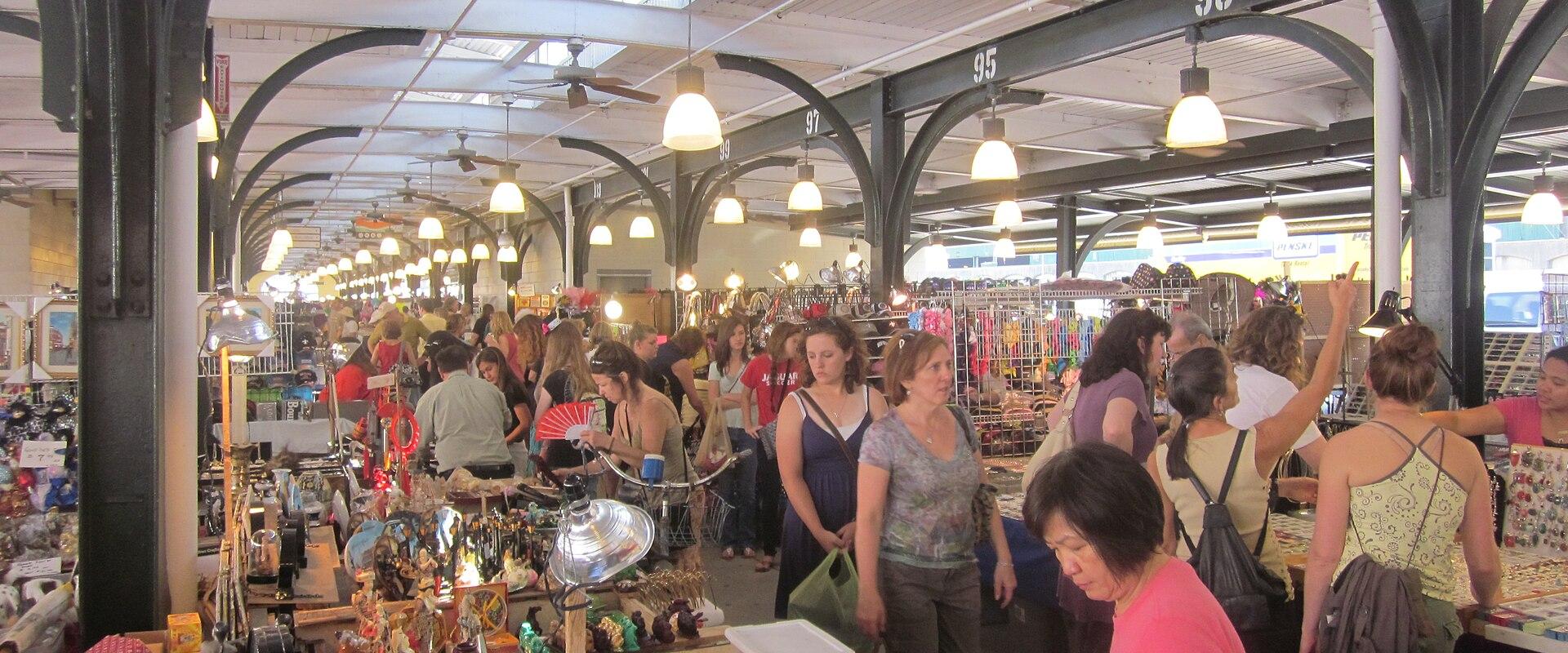 The image shows an indoor flea market with numerous stalls selling antiques and second-hand goods, featuring arched metal beams, hanging lights, and a crowd of shoppers browsing items.