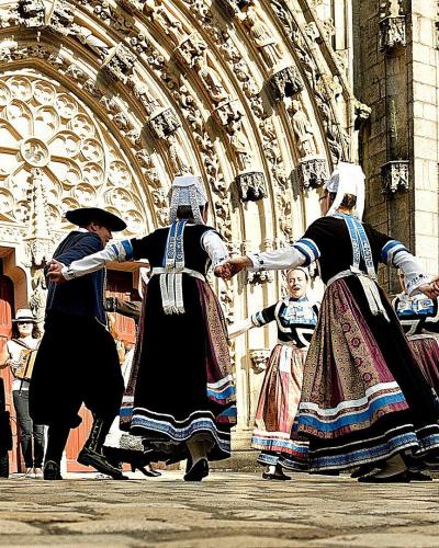 A group of people in traditional Breton folk costumes are dancing in a circle on a paved square, accompanied by musicians with guitar and accordion, directly in front of the ornate Gothic stone facade of a cathedral featuring a large rose window a