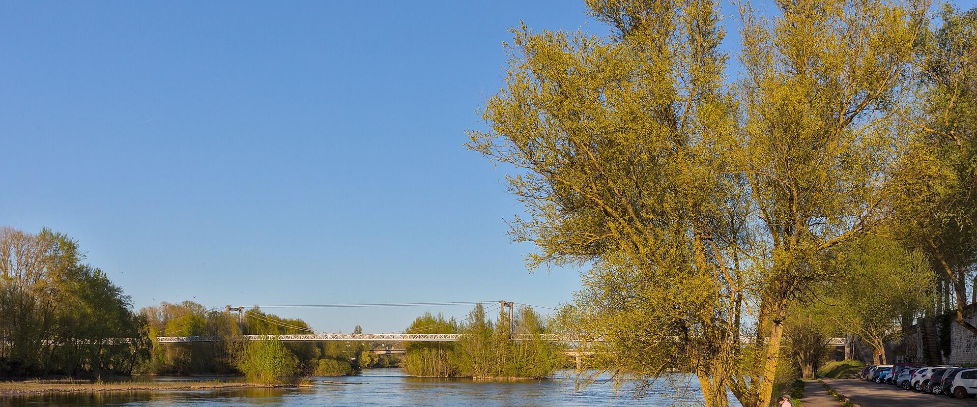 1. A wide river flows under a clear blue sky, with a metal truss bridge in the distance, a tree-lined path on the right bank where cars are parked and people sit, and lush green trees framing the scene.