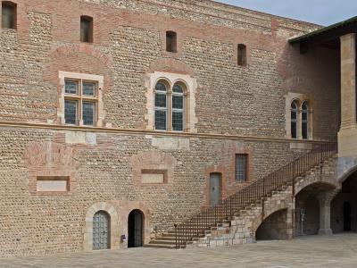 The image shows a large, paved courtyard surrounded by historic stone and brick buildings featuring arched windows, a prominent external stone staircase with a metal railing, and arched ground-floor passageways under an overcast sky.