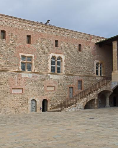 The image shows a large, paved courtyard surrounded by historic stone and brick buildings featuring arched windows, a prominent external stone staircase with a metal railing, and arched ground-floor passageways under an overcast sky.