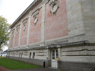 The image shows the exterior of a large, classical-style building with red brick walls, stone quoins and pilasters, decorative oval cartouches, and a white door at ground level, set against a grassy area with trees.