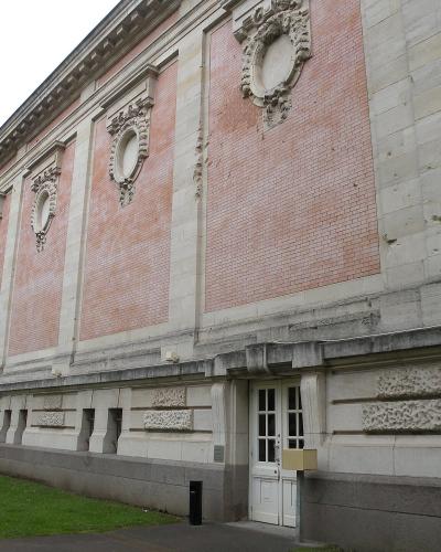The image shows the exterior of a large, classical-style building with red brick walls, stone quoins and pilasters, decorative oval cartouches, and a white door at ground level, set against a grassy area with trees.