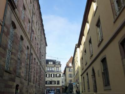 A narrow European street flanked by tall, historic buildings with varied facades, including one with reddish sandstone and another with yellow stucco, under a blue sky; a car is parked in the distance and signage for "Boulangerie" and "Pâtisserie"