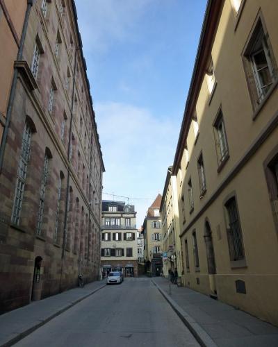 A narrow European street flanked by tall, historic buildings with varied facades, including one with reddish sandstone and another with yellow stucco, under a blue sky; a car is parked in the distance and signage for "Boulangerie" and "Pâtisserie"