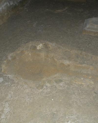 The image shows a dimly lit underground stone floor with a circular stone basin or well-like structure in the center and a rectangular stone block to the right, consistent with an archaeological or historical crypt interior.