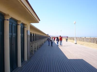 A long wooden boardwalk (plank walkway) runs alongside a sandy beach, featuring a row of distinctive red and blue striped beach chairs and umbrellas in the distance, with people strolling along the path.