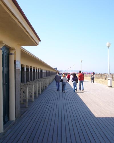 A long wooden boardwalk (plank walkway) runs alongside a sandy beach, featuring a row of distinctive red and blue striped beach chairs and umbrellas in the distance, with people strolling along the path.