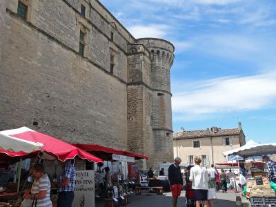 A low-angle outdoor shot of a large, light-colored stone fortress with a prominent round tower, set against a blue sky, with market stalls featuring red and white awnings and shoppers in the foreground.