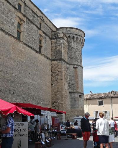 A low-angle outdoor shot of a large, light-colored stone fortress with a prominent round tower, set against a blue sky, with market stalls featuring red and white awnings and shoppers in the foreground.