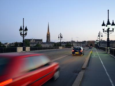 A view from a bridge at dusk showing tram tracks, a dedicated bike lane, ornate street lamps, and a prominent Gothic church spire in the background.