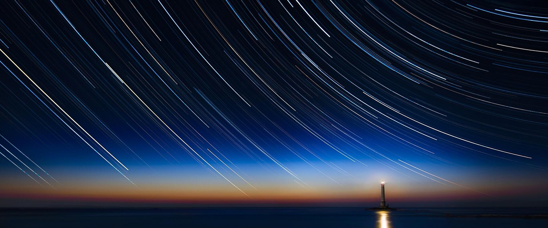 A long-exposure night photograph showing star trails arching across the sky above a solitary lighthouse on a rocky outcrop over water, with a faint glow on the horizon.