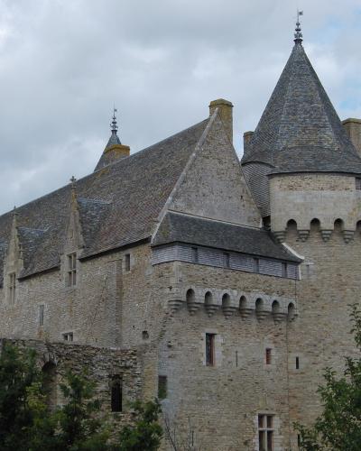 A stone medieval castle featuring a large round tower with a conical slate roof, decorative gables, and crenellations, set against a cloudy sky with green foliage in the foreground.