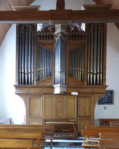 The image shows the interior of a church, focusing on a large wooden pipe organ with gold detailing, set against white walls and a wooden ceiling, with rows of wooden pews in the foreground.