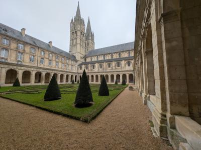 A stone cloister courtyard with manicured boxwood hedges and conical topiary trees, surrounded by arched walkways and a large Gothic-Romanesque church tower in the background under an overcast sky.