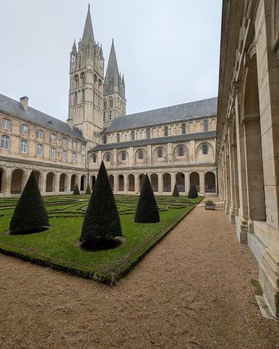 A stone cloister courtyard with manicured boxwood hedges and conical topiary trees, surrounded by arched walkways and a large Gothic-Romanesque church tower in the background under an overcast sky.