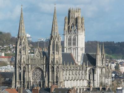 A large Gothic church with two tall spires on the left, a central rose window, and a massive ornate tower on the right, set against a backdrop of urban buildings and hills under a partly cloudy sky.