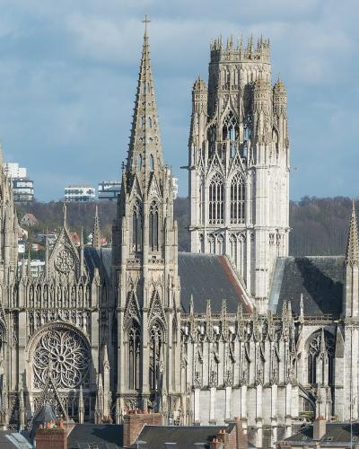 A large Gothic church with two tall spires on the left, a central rose window, and a massive ornate tower on the right, set against a backdrop of urban buildings and hills under a partly cloudy sky.