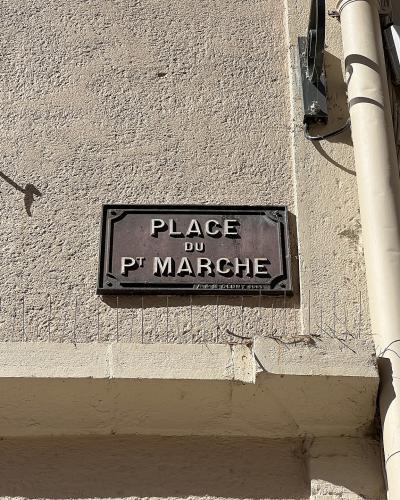 A close-up photograph of a dark, rectangular street sign mounted on a textured beige stucco wall, reading "PLACE DU PT MARCHE" with smaller text below indicating "FABR. DE CLUNY 2005".