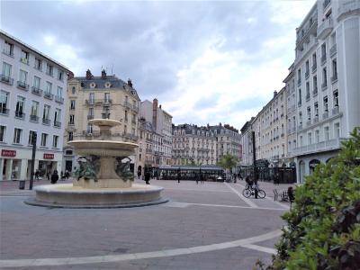 A wide-angle photograph of a large, paved European city square featuring a prominent multi-tiered stone fountain with bronze sculptures in the foreground, surrounded by classical-style buildings and a few pedestrians.