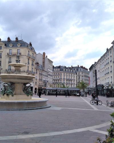 A wide-angle photograph of a large, paved European city square featuring a prominent multi-tiered stone fountain with bronze sculptures in the foreground, surrounded by classical-style buildings and a few pedestrians.