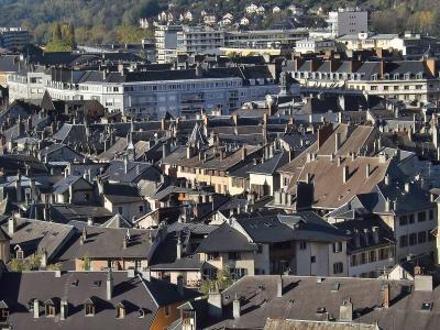 A high-angle view of a dense European cityscape featuring numerous slate and tile rooftops, chimneys, and historic buildings, with a modern white building and a hillside in the background.